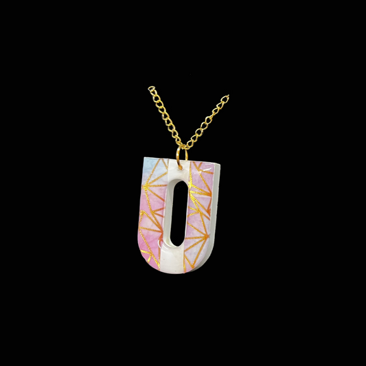 Ge din stil en personlig touch med detta unika bokstavshalsband i formen av U. Hänget har mjuka pastelltoner av rosa, blått och vitt, dekorerat med eleganta geometriska mönster i skimrande guld. Ett trendigt och tidlöst smycke att bära till vardags eller som en färgklick till festklädseln.